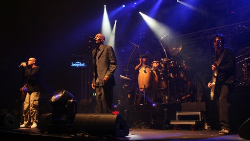 Großaufnahme von Faithless: in der Mitte Maxi Jazz, umrahmt von Background-Musikern, hinten leuchtet das helle Holz der Drums.