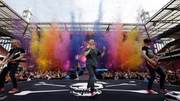 Der Gitarrist von Kasalla (l.), der Sänger (M.) und der Bassist (r.) stehen auf der Bühne, im Hintergrund das Publikum im Kölner Stadion und mehrere Farbbomben, die in diesem Moment vor der Bühne ein buntes Bild verursachen.