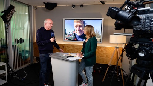 Gespräch zweier Personen an einem Stehtisch im Fernsehstudio, im Hintergrund ist auf dem Wandmonitor eine weitere Person zu sehen