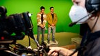 Im Hintergrund stehen zwei Schüler als Moderatoren vor einer grünen Wand, im Vordergrund bedient ein:e weitere:r eine Fernsehkamera