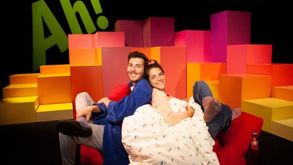 Die Moderatoren Tarkan Bagci und Clarissa Corrêa da Silva sitzen auf einer roten Couch, zugedeckt mit einer Kinderbettdecke