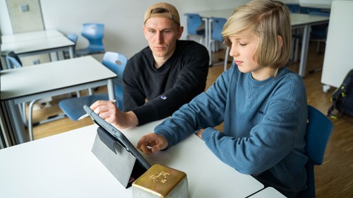 Ein Schüler und ein Mann sitzen vor einem Tablet mit einem goldenen Stolperstein.