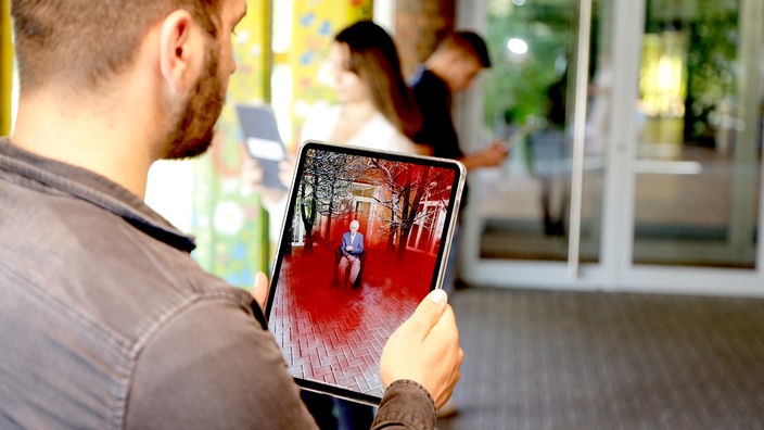 Schüler hält ein Tabletcomputer vor sich, auf dem die WDR History App läuft. Im Hintergrund stehen zwei weitere Schüler und betrachten ihre Tablets.