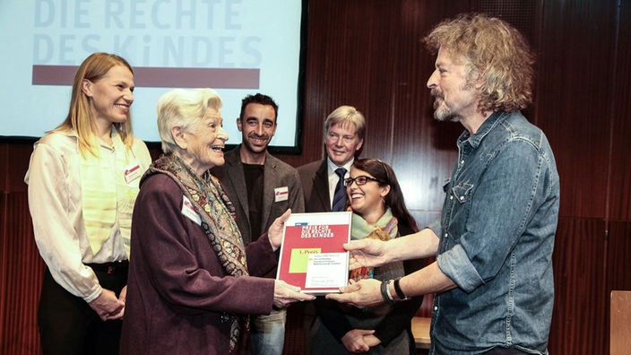 Menschen auf der Bühne,  Schirmherr Wolfang Niedecken überreicht den 1. Preis an "Andehri Hilfe" aus Bonn