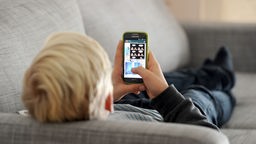 Das Bild zeigt ein Kind, das mit einem Smartphone auf der Couch liegt.