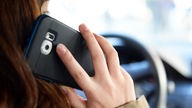 Das Bild zeigt eine Dame, die mit dem Handy am Ohr hinter dem Steuer eines Autos sitzt.