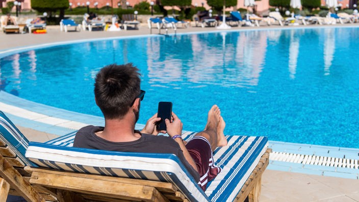 Ein Mann sitzt im Urlaub mit seinem Handy am Pool.