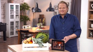 Björn Freitag in der Küche neben einer Kiste mit Gemüse und einem Tablet in der Hand.