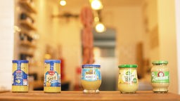 fünf verschiedene Marken Senf nebeneinander auf einem Holztisch präsentiert