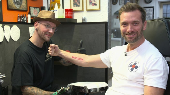 Das Bild zeigt Daniel Aßmann mit einem Tattoo-Artist in einem Tattoo-Studio.