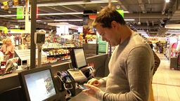 Ein Mann steht an einer SB-Kasse und bereitet sein Smartphone fürs mobile Bezahlen vor