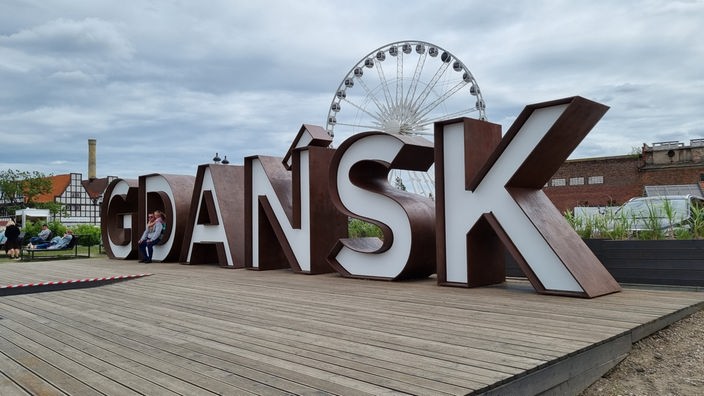 Das Foto zeigt den Schriftzug der Stadt Gdansk (Danzig) als dreidimensionales Objekt. Im Leerraum des Buchstaben 'D'sitzt eine Person. Im Hintergrund ein Riesenrad.