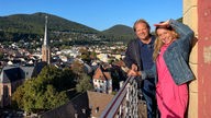 Das Bild zeigt Björn Freitag (l.) mit Tamina Kallert (r.) in der Pfalz auf einem Balkon stehend.