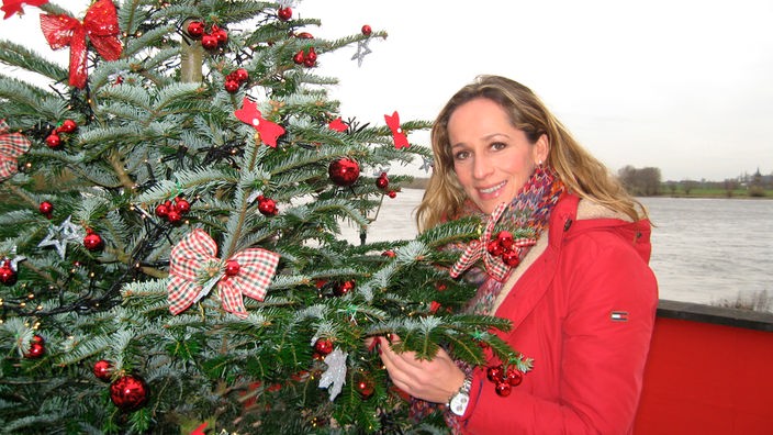 Die Moderatorin Tamina Kallert steht neben einem rot geschmückten Weihnachtsbaum.