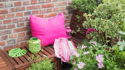Das Bild zeigt einen Balkon mit Bank, pinkem Kissen, einer Decke, einer Laterne und diversen Topfpflanzen.