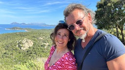 Das Bild zeigt Britta (links) und Andreas (rechts) mit der sommerlichen Landschaft Sardiniens im Hintergrund.