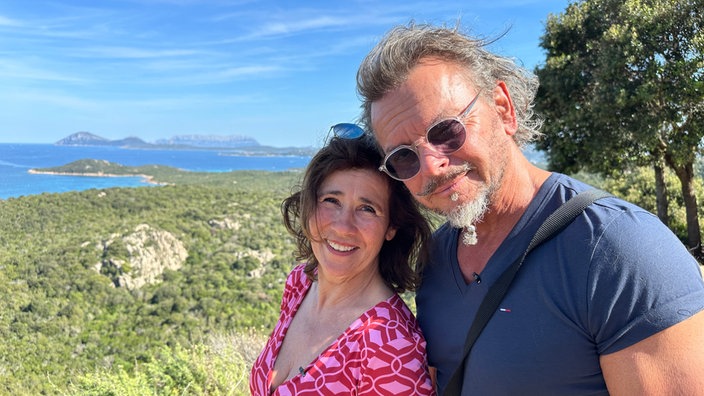 Das Bild zeigt Britta (links) und Andreas (rechts) mit der sommerlichen Landschaft Sardiniens im Hintergrund.