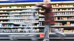 Das Bild zeigt eine Kundin, die mit einem Einkaufswagen durch den Supermarkt geht.