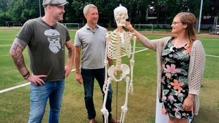 Das Bild zeigt Dr. Esser auf einem Sportplatz mit Zusammen mit dem Orthopäden Prof. Christopher Niedhart und Bürokauffrau Claudia und einem Skelett.