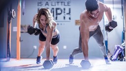 Das Bild zeigt einen Mann und eine Frau beim Sport mit Hanteln im Fitnessstudio.