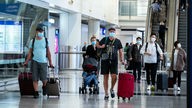 Das Bild zeigt Touristen mit Koffern und Mundschutz am Flughafen.