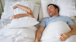 Mann schnarcht und Frau hält sich Kissen vor das Gesicht.