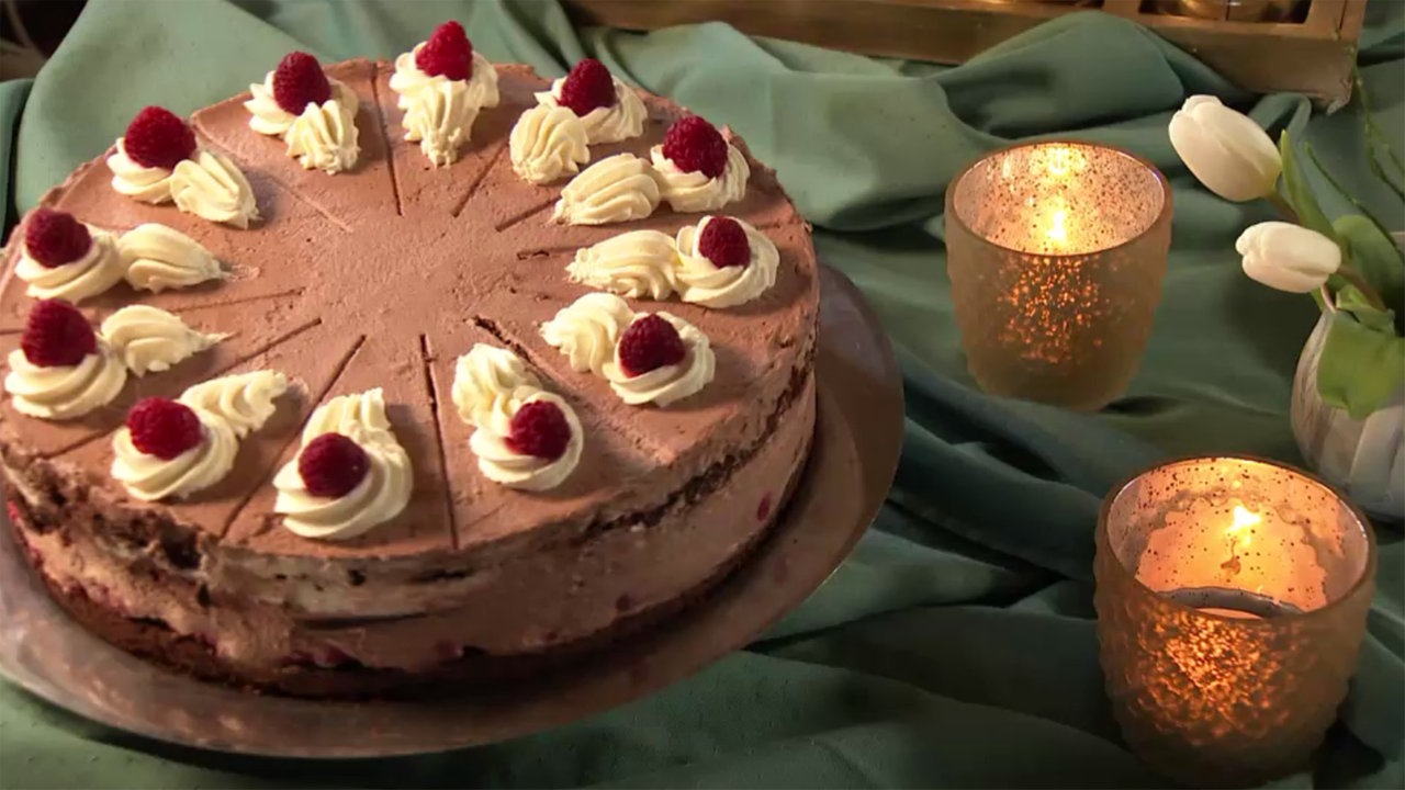 Auf einem grünen Tuch steht links eine Himbeer-Schoko-Torte mit Himbeeren auf den Sahne-Tupfern, rechts Teekerzen.