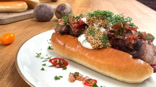 Das Bild zeigt einen Hotdog, gefüllt mit Schaschlik und verschiedenen Soßen, garniert mit Dill und Chili.