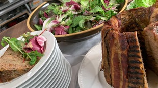 Angeschnittener Guglhupf auf einer Platte, daneben ein Teller mit einem Stück Guglhupf und Salat angerichtet, im Hintergrund eine Schüssel mit buntem Blattsalat 