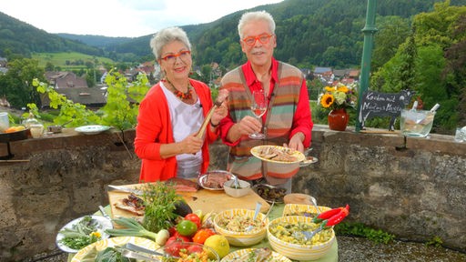 Martina und Moritz stehen auf einer Terrasse hinter einem Tisch mit verschiedenen Gerichten und Zutaten