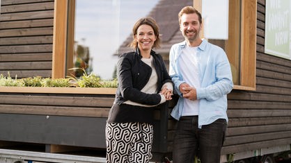 Das Bild zeigt die Moderatoren Anna Planken und Daniel Aßmann vor einem Tiny House auf Rädern.