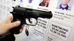 Eine mit der Mordwaffe baugleiche Pistole wird im Polizeipräsidium in Dortmund vor eine Bilderwand mit den Porträts von Opfern einer deutschlandweiten Mordserie gehalten