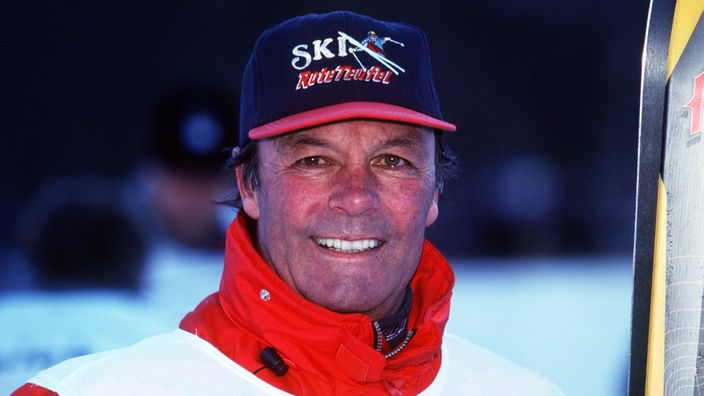 Toni Sailer in Anorak mit Kappe und Skiern lächelt in die Kamera (um 1985)