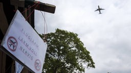 Am Himmel ist ein Flugzeug zu sehen, an einem Schuppen ist ein Schild mit dem Text: "Fluglärm, Lärm voll nervig" angebracht.