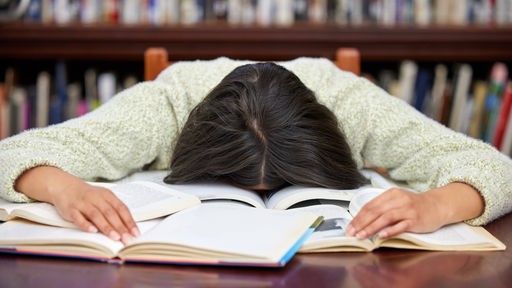 Eine Frau legt ihren Kopf erschöpft auf einem aufgeschlagenen Buch ab.
