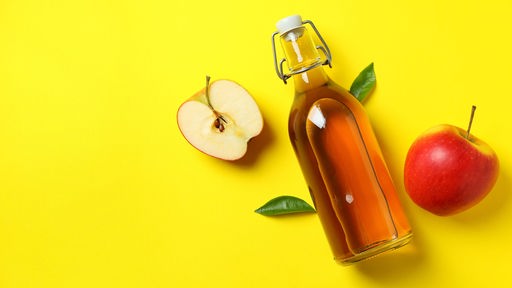 Eine Bügelflasche mit Apfelessig liegt neben Äpfeln auf gelbem Grund.