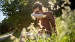 Ein Mann niest in einem Park auf einer Wiese in seine Armbeuge.
